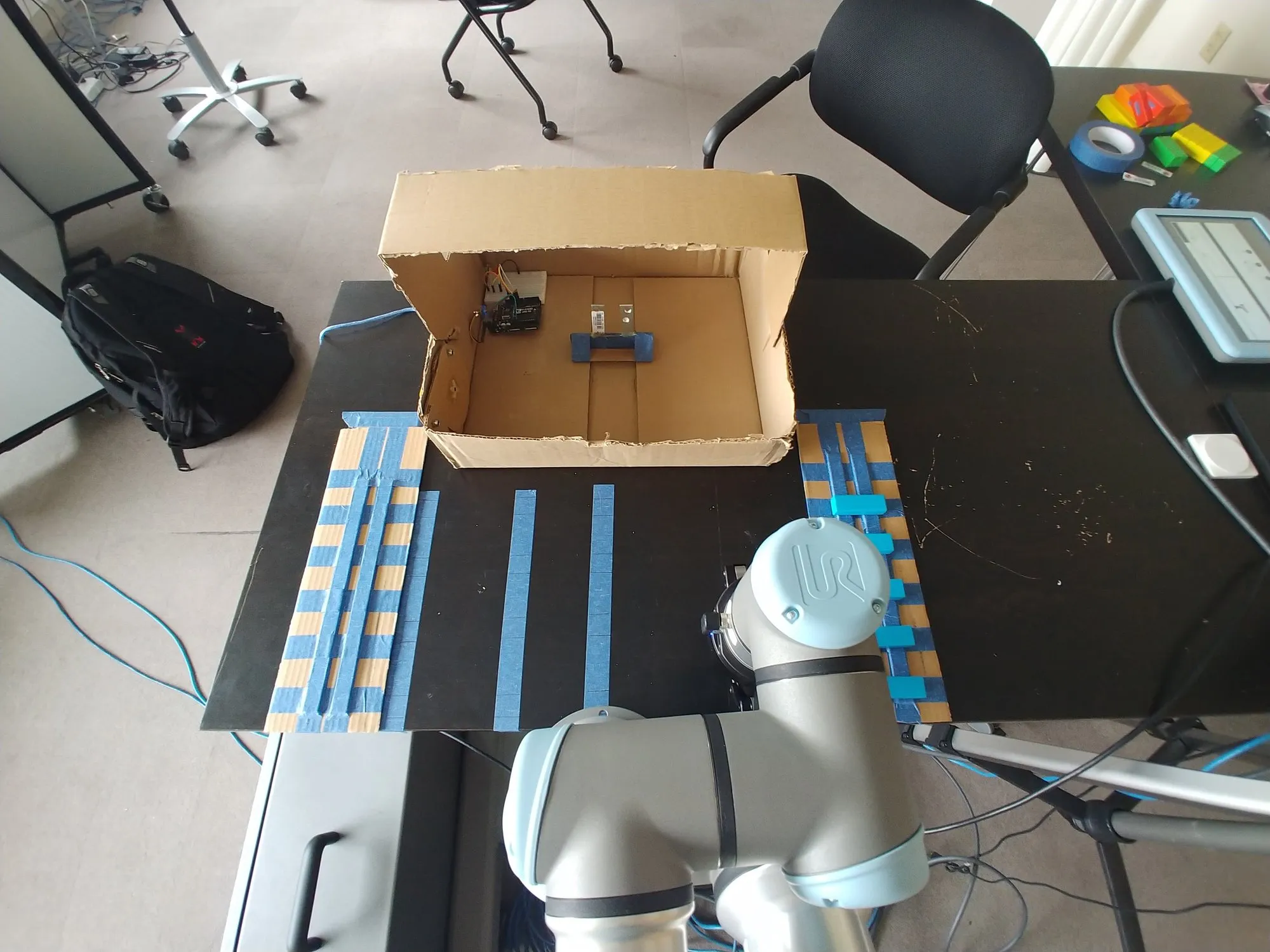 The box at the far end of the table with the taped block grid in front of it, the arm at the near edge.
