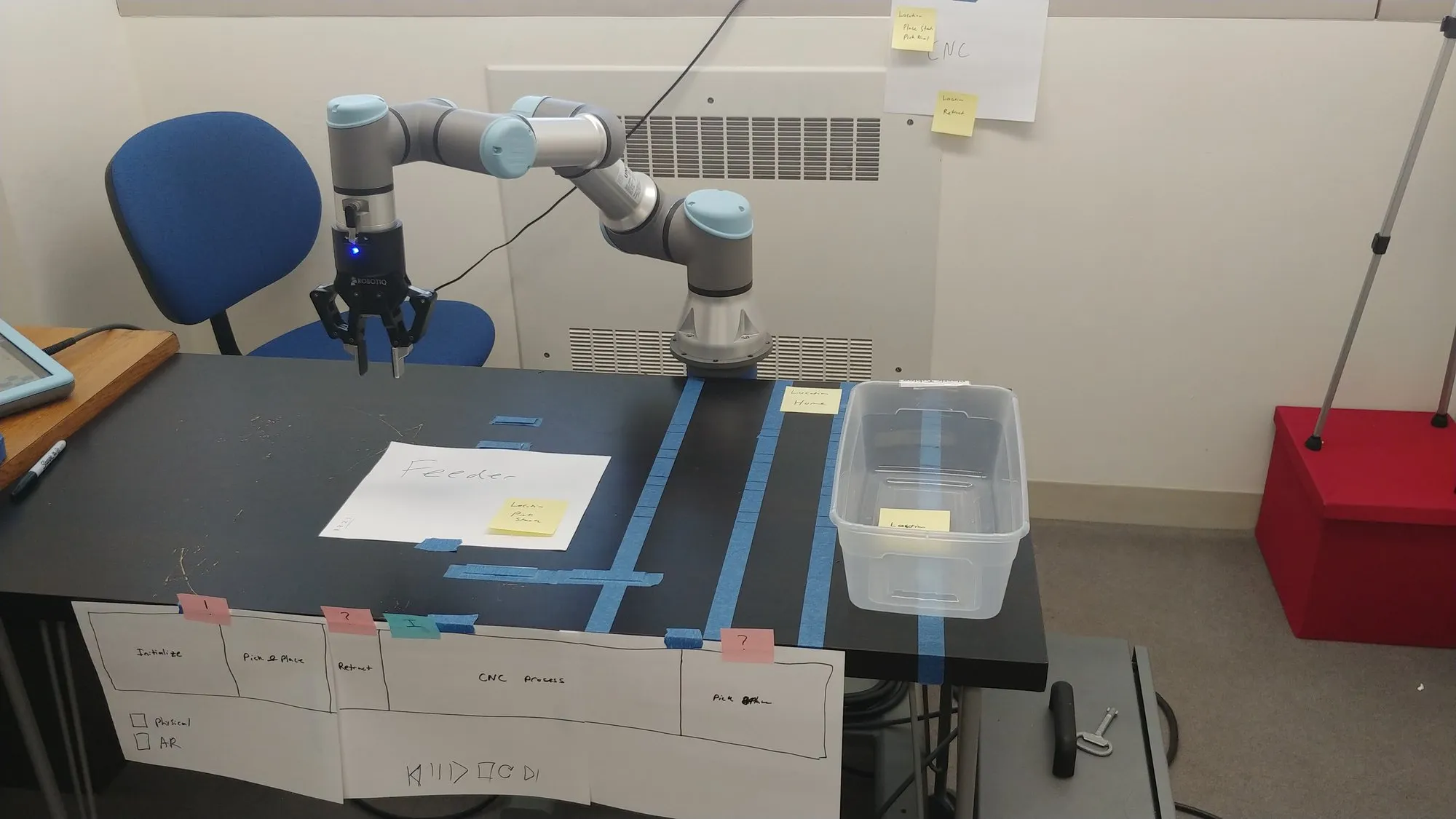 The February 2020 workspace: the UR3e over the taped table with a clear container and a paper sheet, task cards along the front edge.