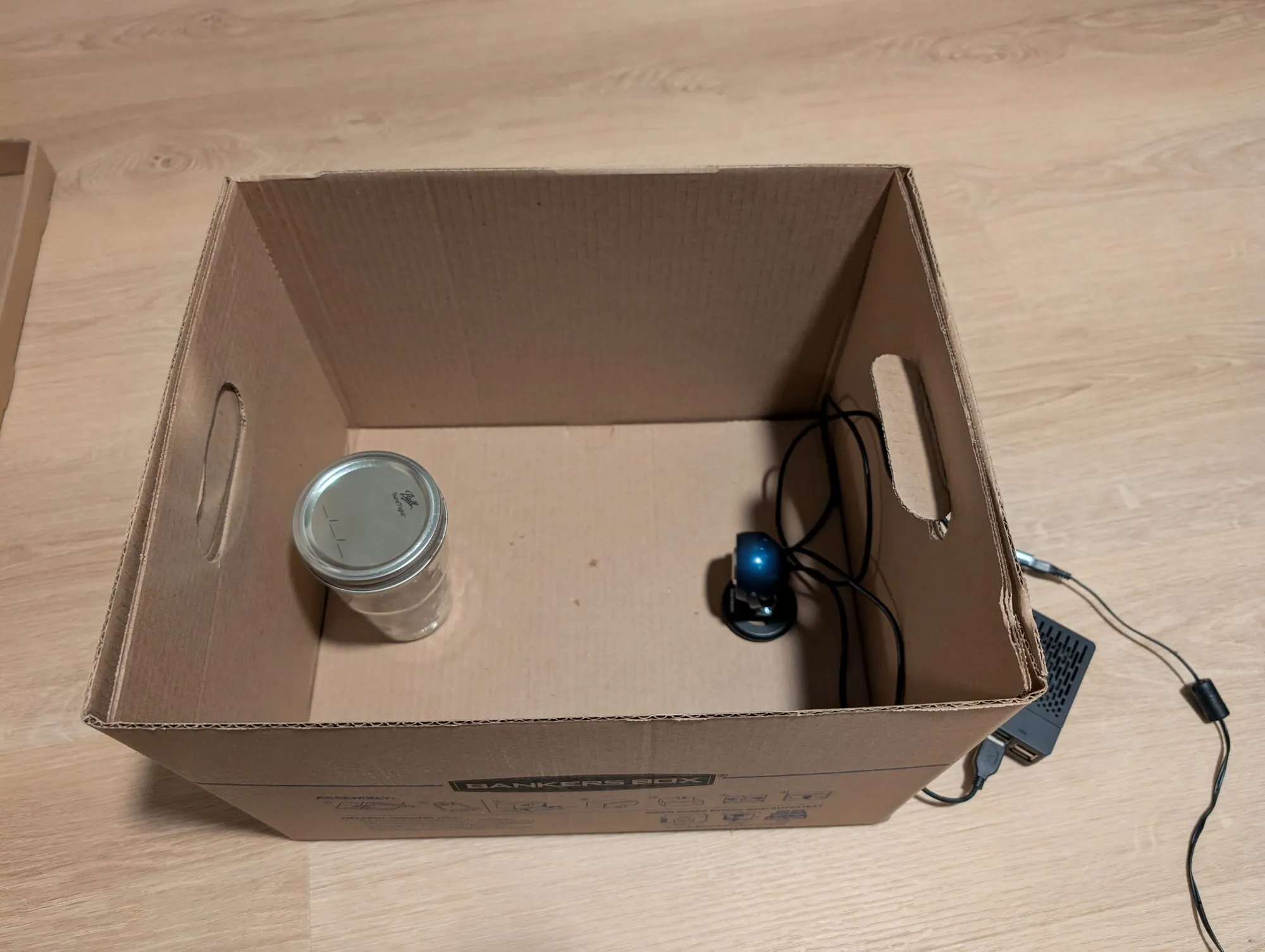 An open Bankers Box from above: an empty mason jar at the left, a blue USB webcam on a round base at the right pointed at it, its cable running out a handle hole to a black Raspberry Pi case outside the box.