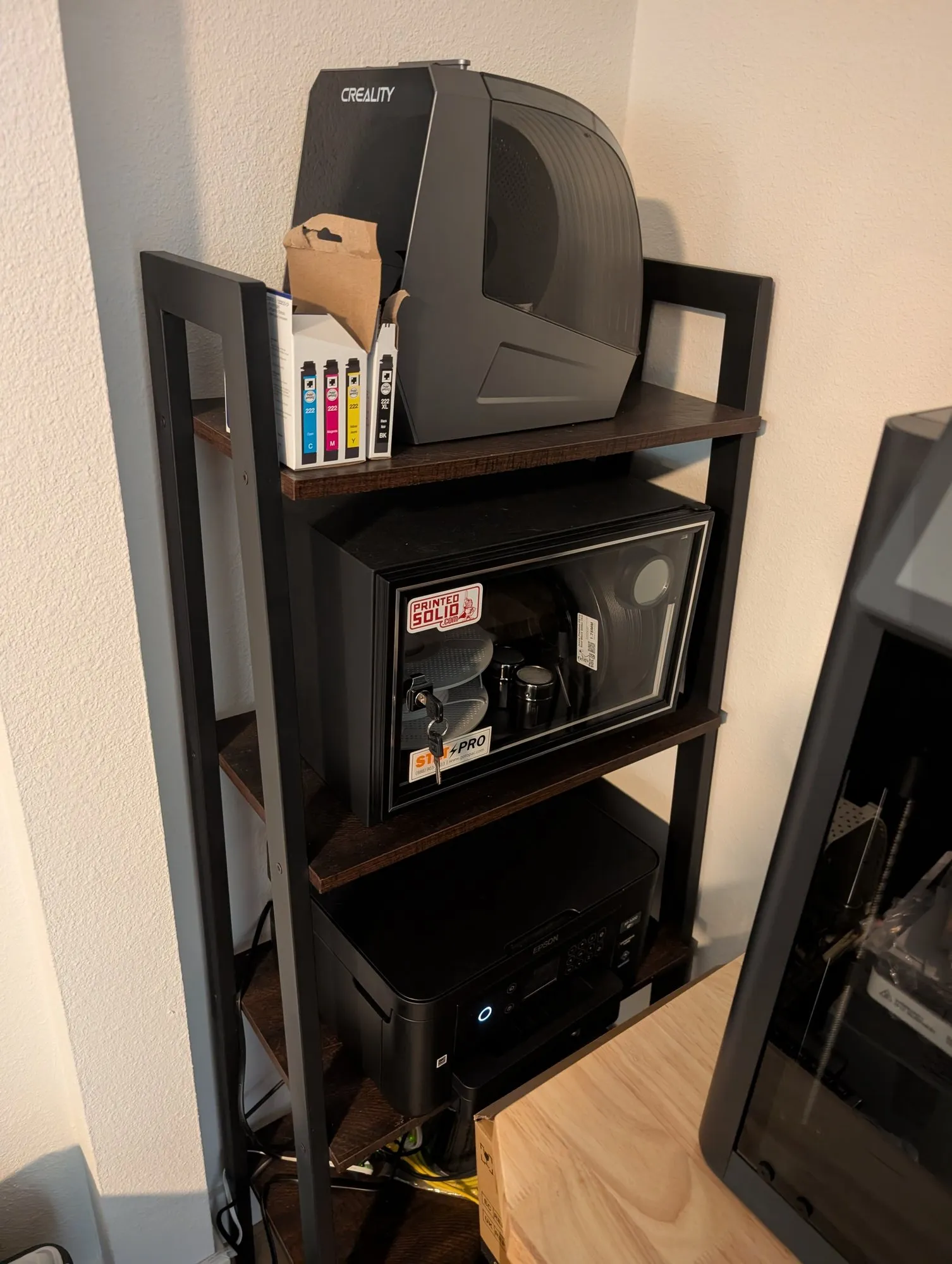 A black shelving unit in a corner: a Creality filament dryer on the top shelf beside boxes of ink cartridges, a locked glass-front dry cabinet holding filament spools on the middle shelf, and an Epson printer on the bottom shelf.