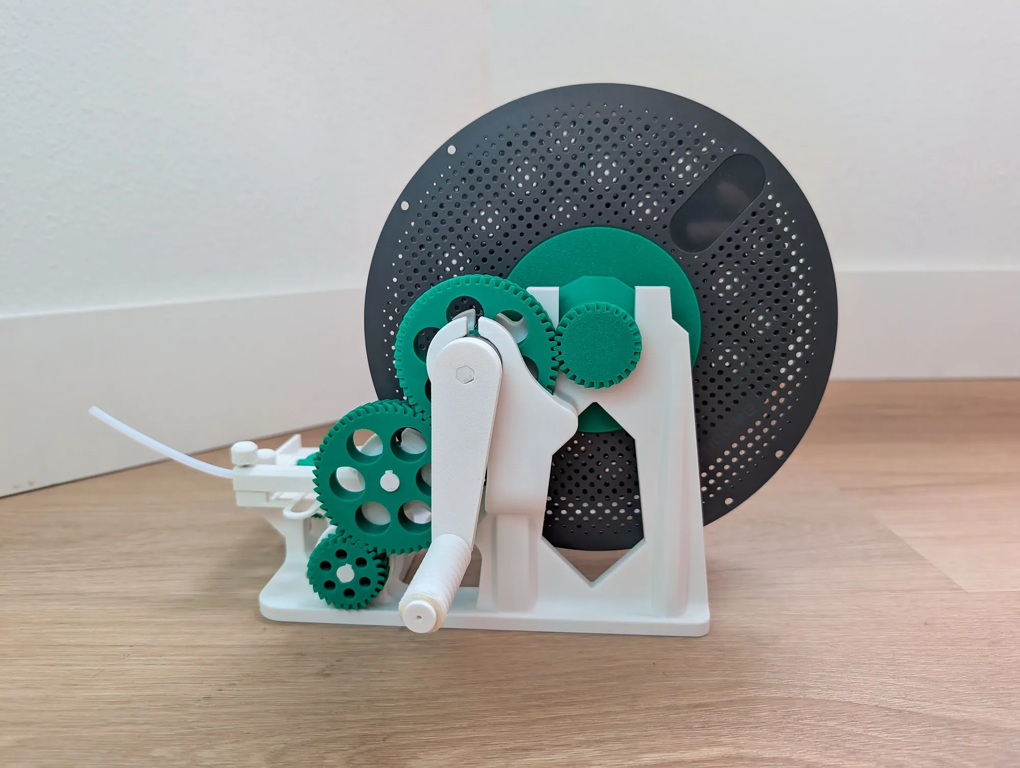 A 3D printed filament spooler on a wood floor, green gears and a white crank arm mounted beside a gray plastic spool.