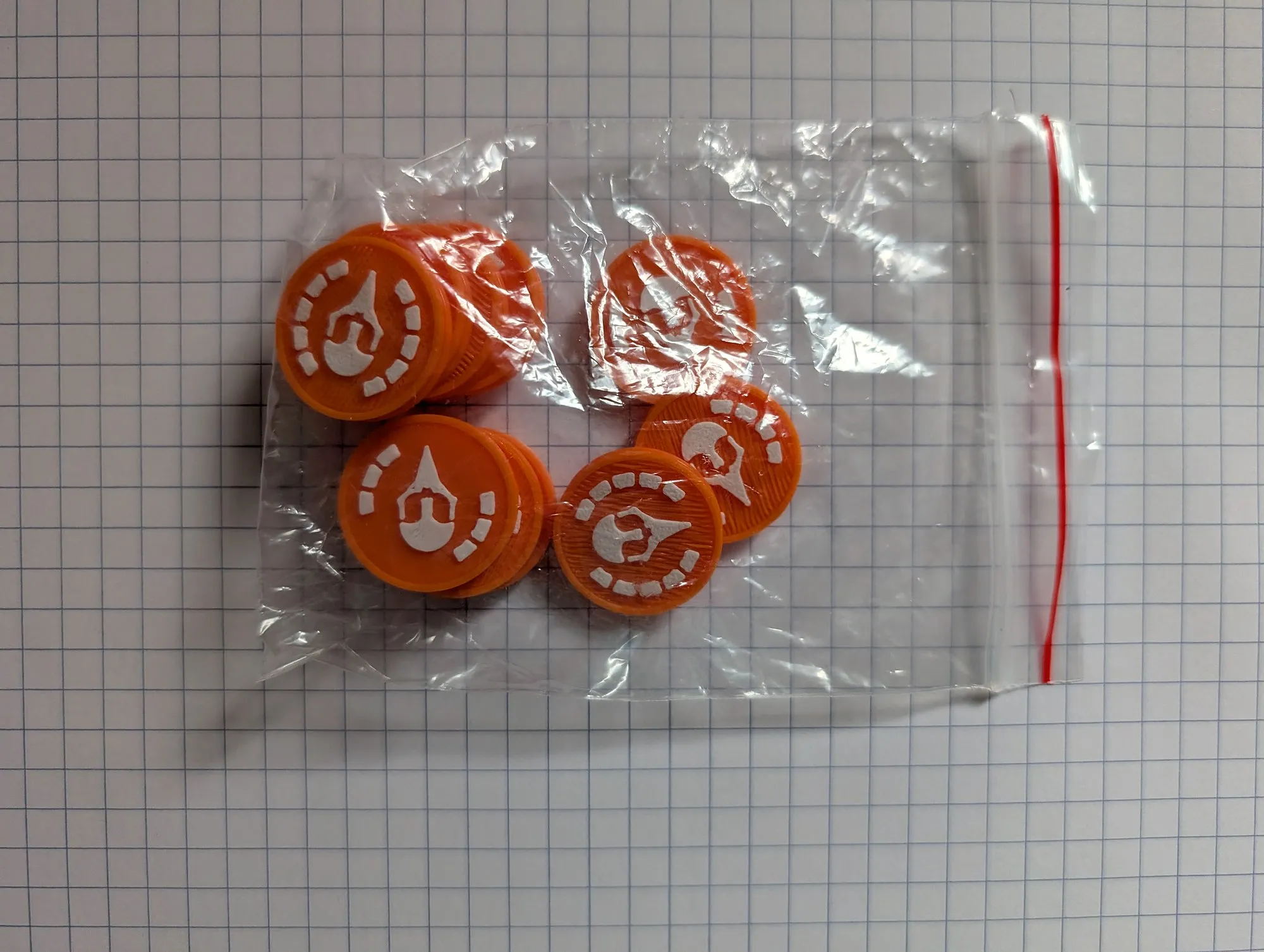 A small zip bag on grid paper holding a handful of orange 3D printed tokens with white painted symbols on top.