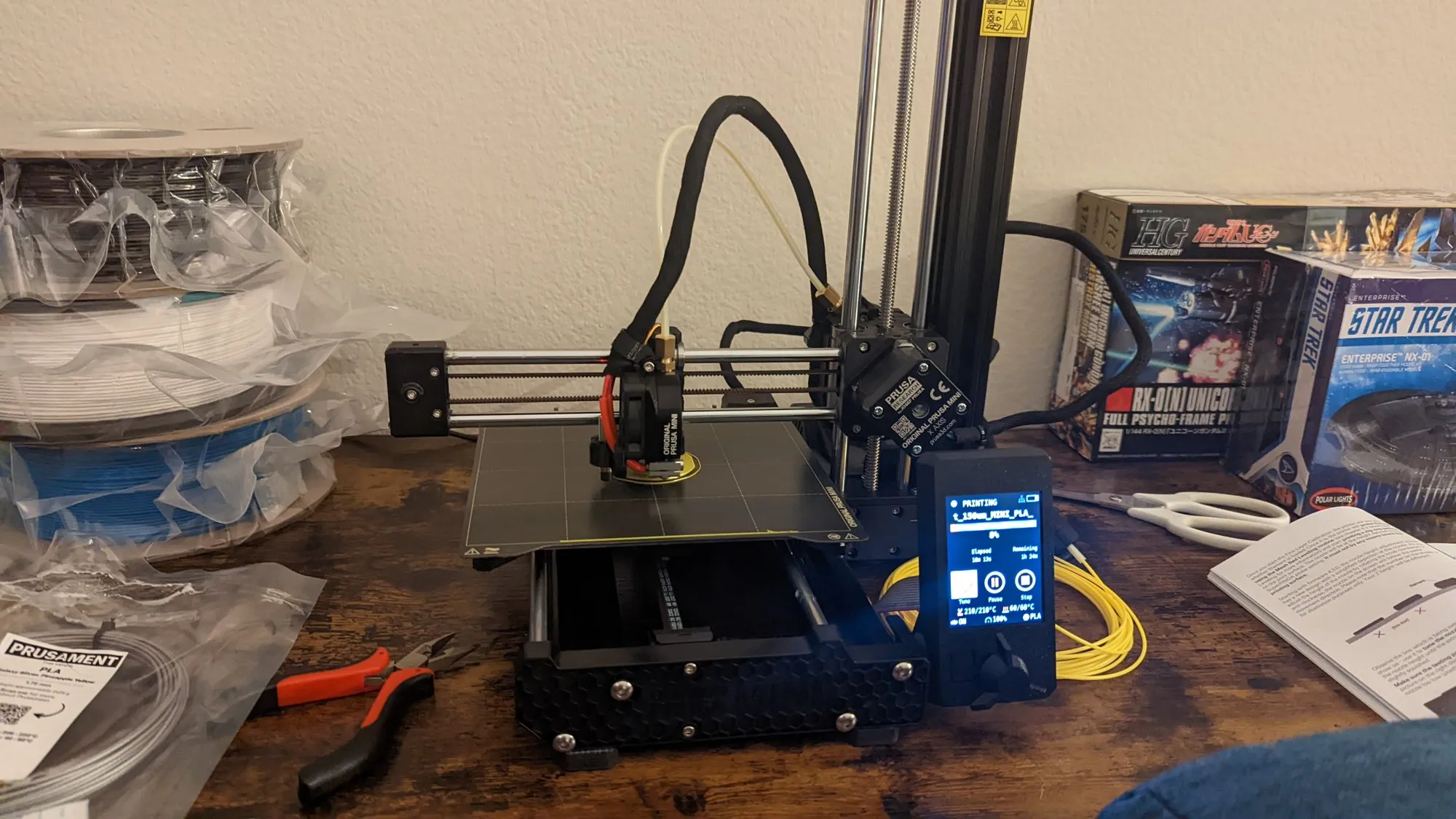 The Prusa Mini on a wooden desk mid-print, filament spools and a pair of pliers beside it, a Gunpla box behind.