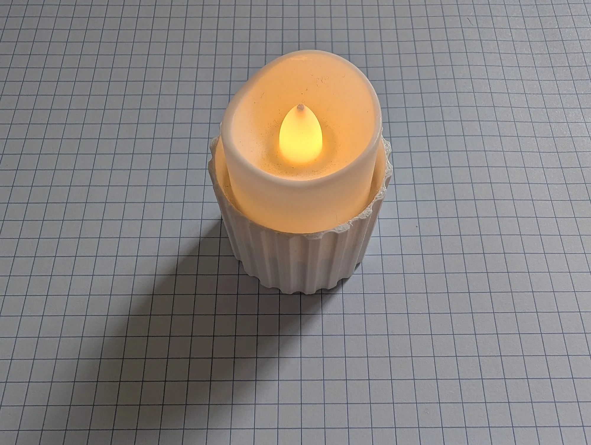 A white fluted 3D printed candle holder on grid paper with a lit LED tealight sitting inside it.
