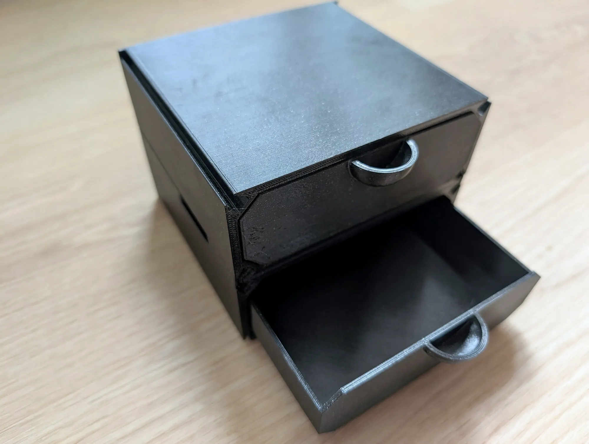 A small black 3D printed box with two drawers on a wood floor, the bottom drawer pulled open.
