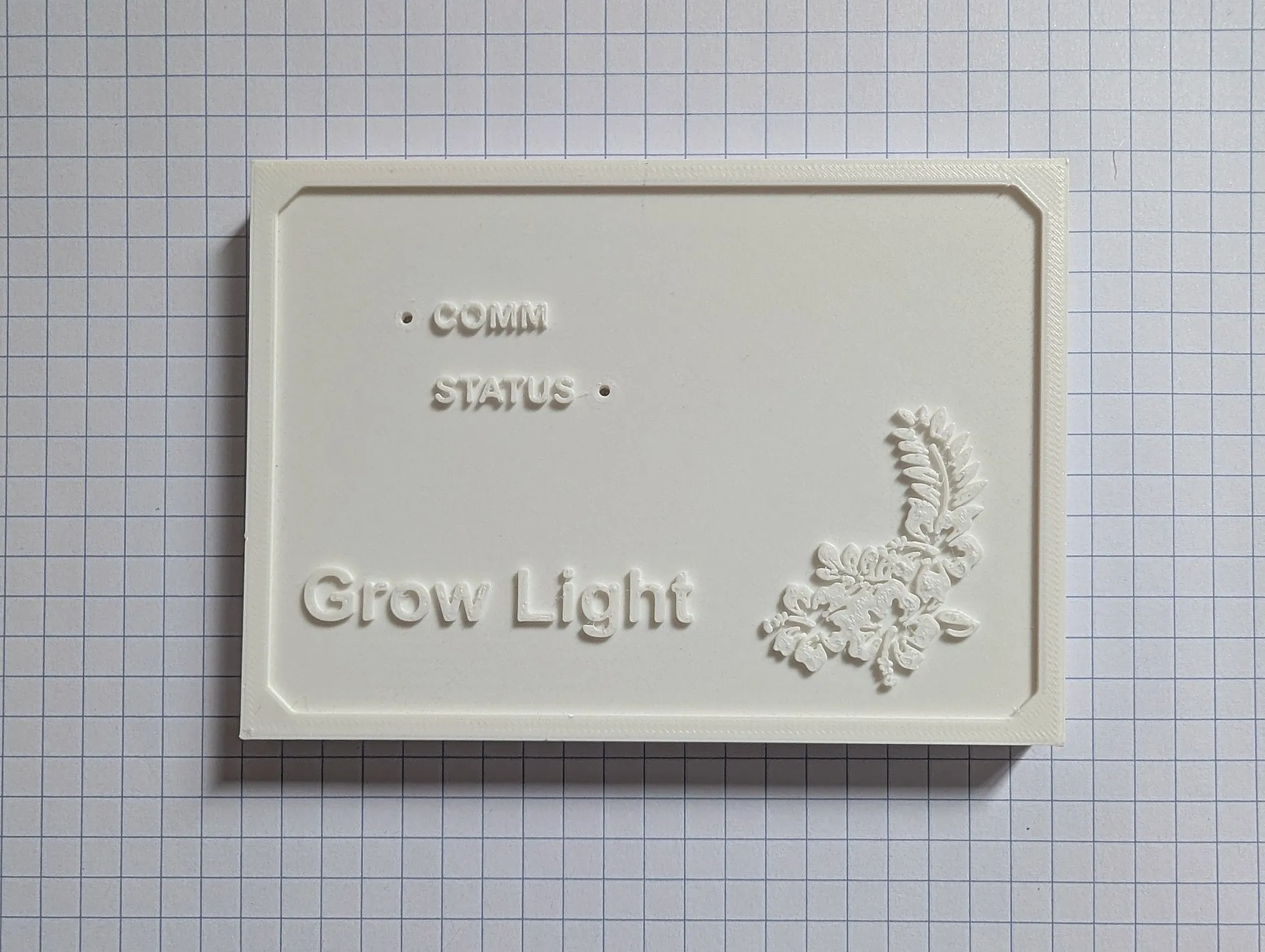 A white 3D printed faceplate on grid paper reading Grow Light, with COMM and STATUS labels and a raised flower in one corner.