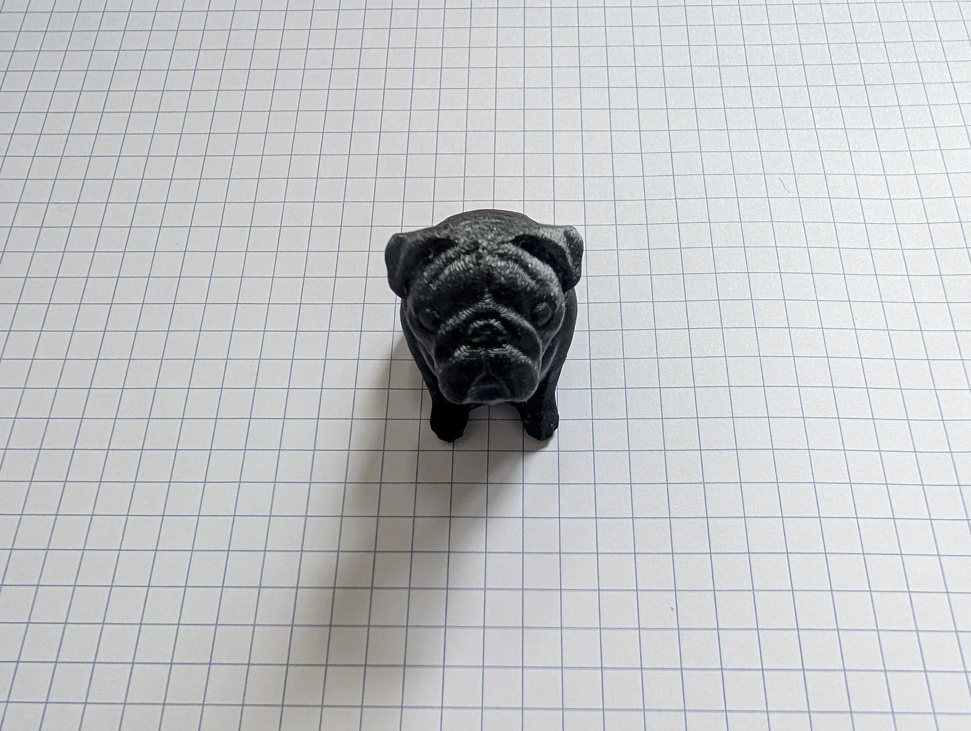 A small black 3D printed pug figurine sitting on grid paper, facing the camera.