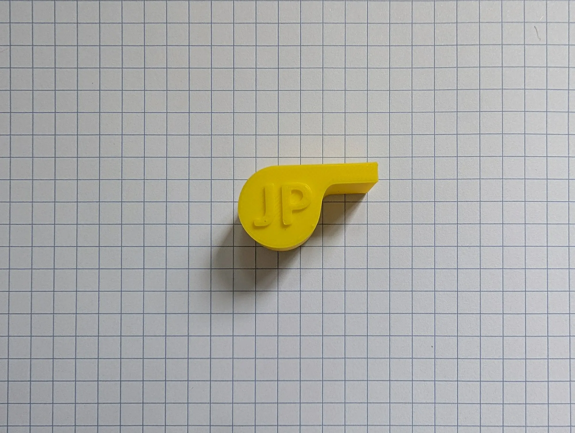 A yellow 3D printed whistle with the letters JP raised on its side, lying on grid paper.