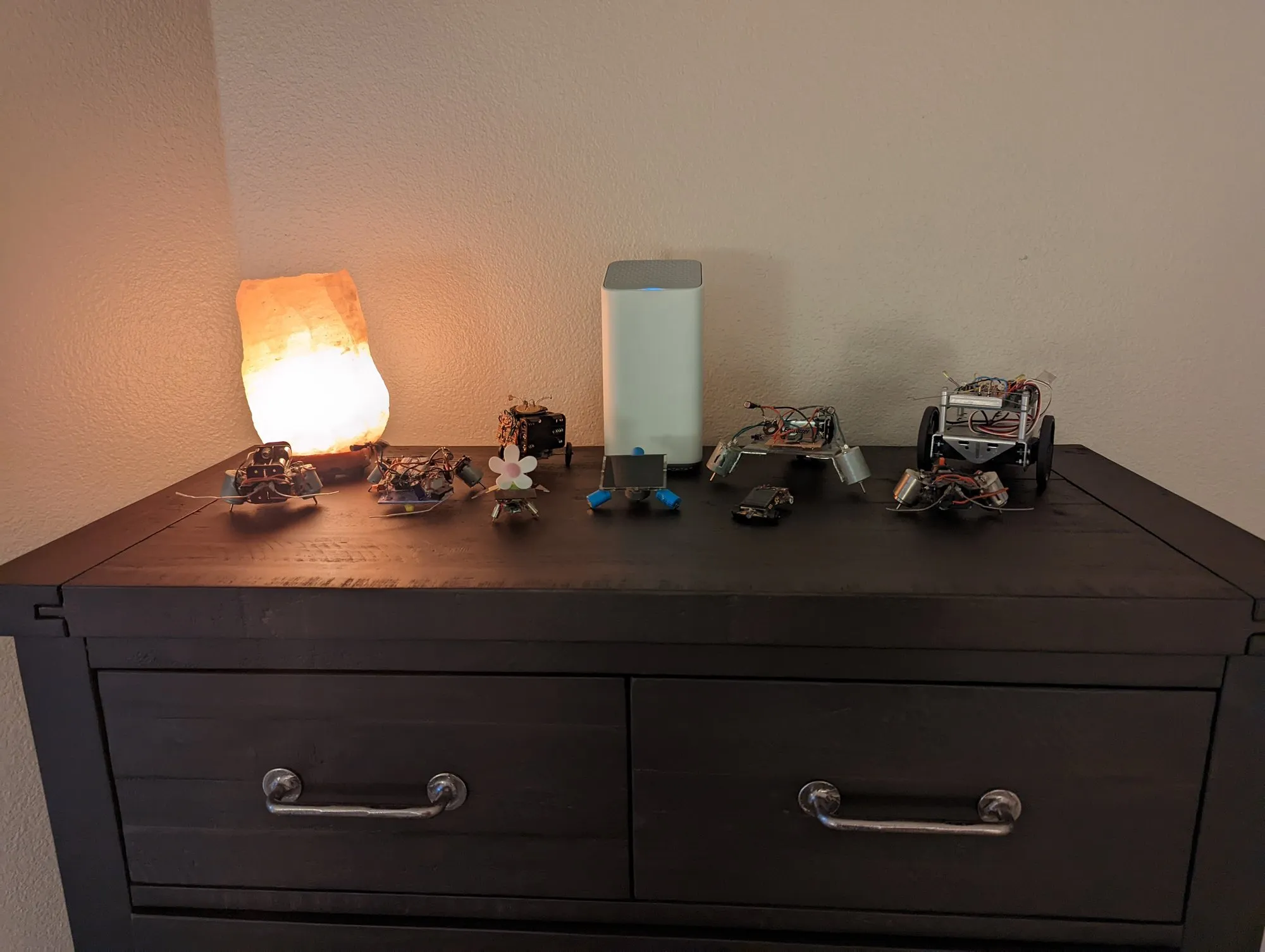 The restored robots lined up on a dark wood dresser in February 2022, a salt lamp glowing at the left end and a Wi-Fi router in the middle: the BEAM bots, Triclops, and the BOE-Bot on the right.