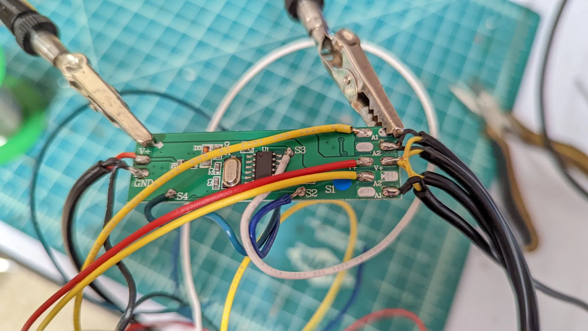 The controller PCB held in two alligator clips over the cutting mat, red, yellow, blue, and white hookup wire freshly soldered to its pads.