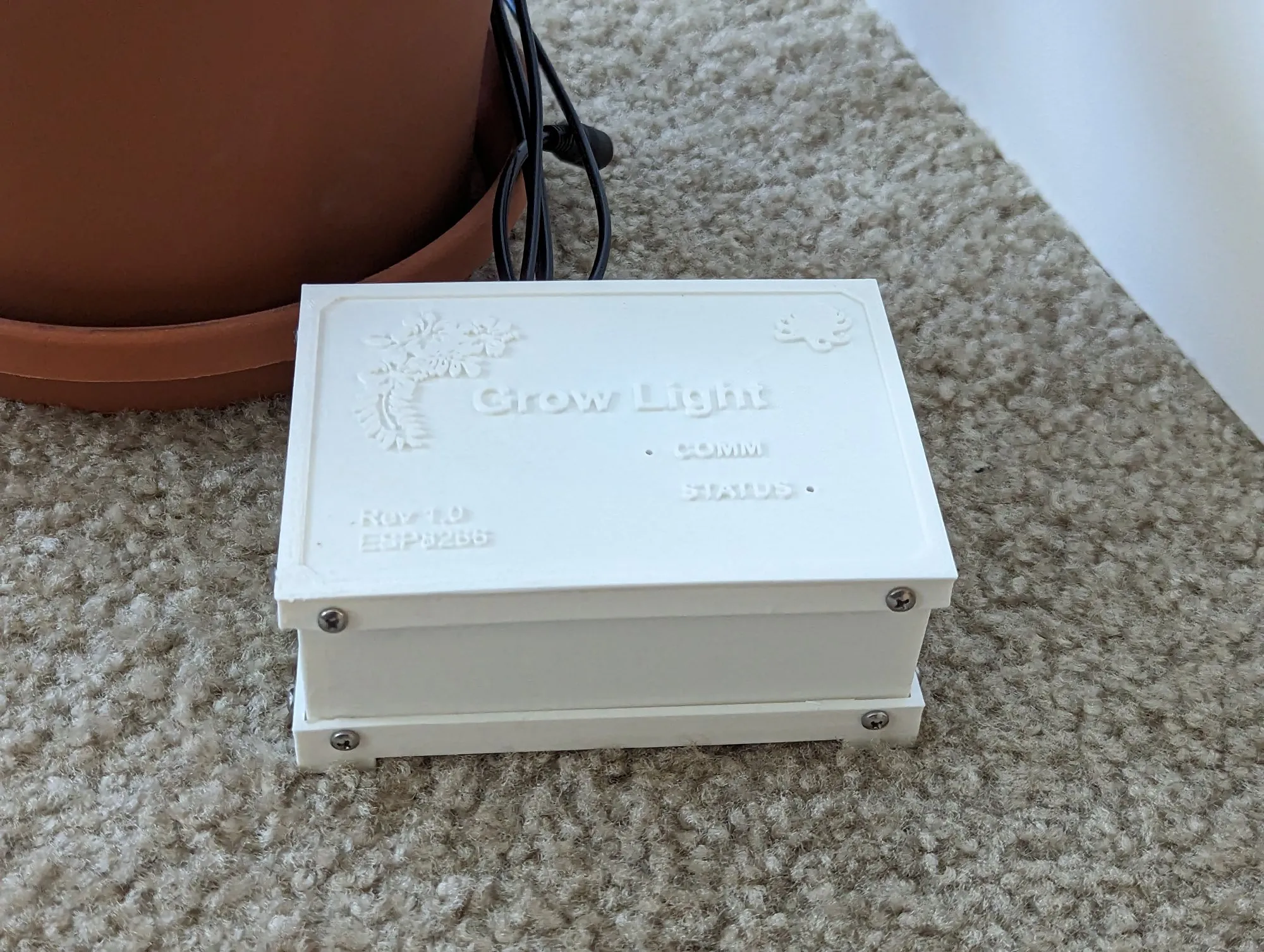 The closed white enclosure on carpet, lid on, next to the base of a terracotta pot.