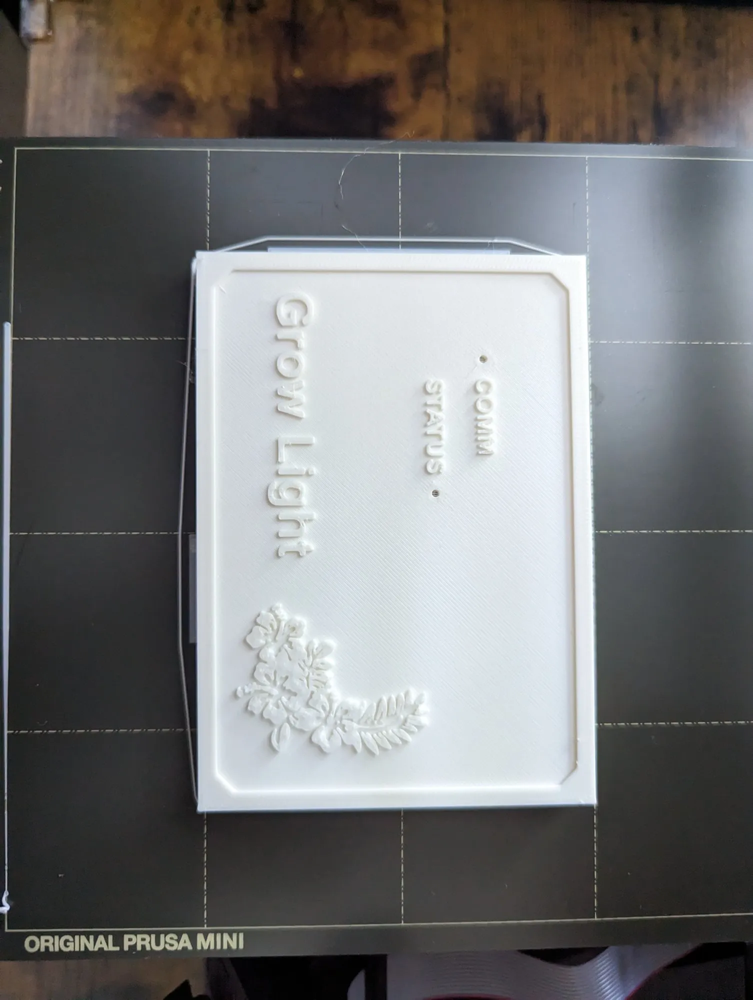 The finished lid on the print bed, Grow Light embossed across it with a small plant logo and two smaller labels reading COMM and STATUS.