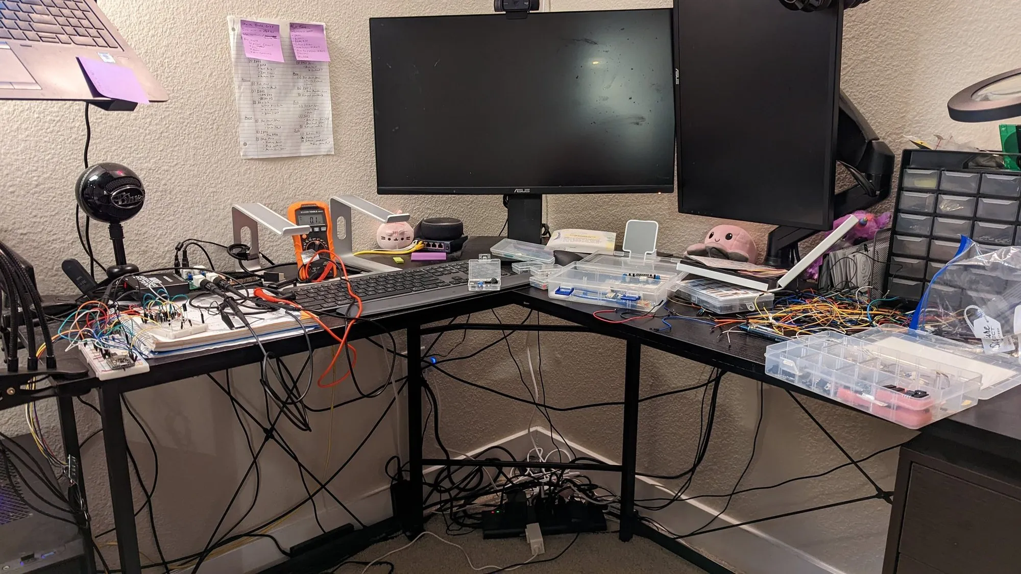 The electronics bench: two monitors, a multimeter, a keyboard, breadboards and parts organizers, a microphone, and a sheet of sticky notes on the wall.