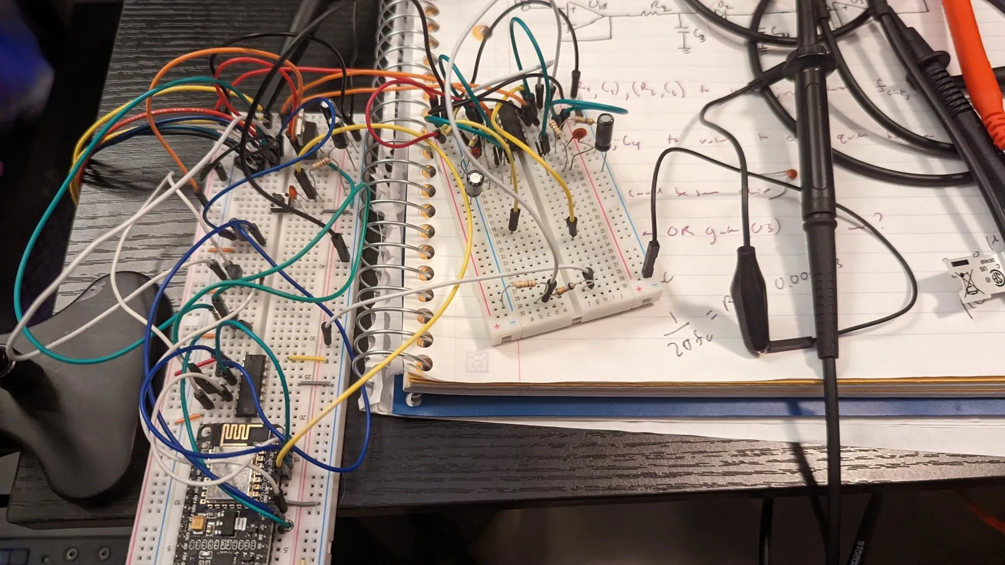 Two breadboards on a desk: a NodeMCU on the left one with jumpers running to op-amp and logic chips on the right one, a spiral notebook underneath with handwritten filter equations and an oscilloscope probe clipped in.