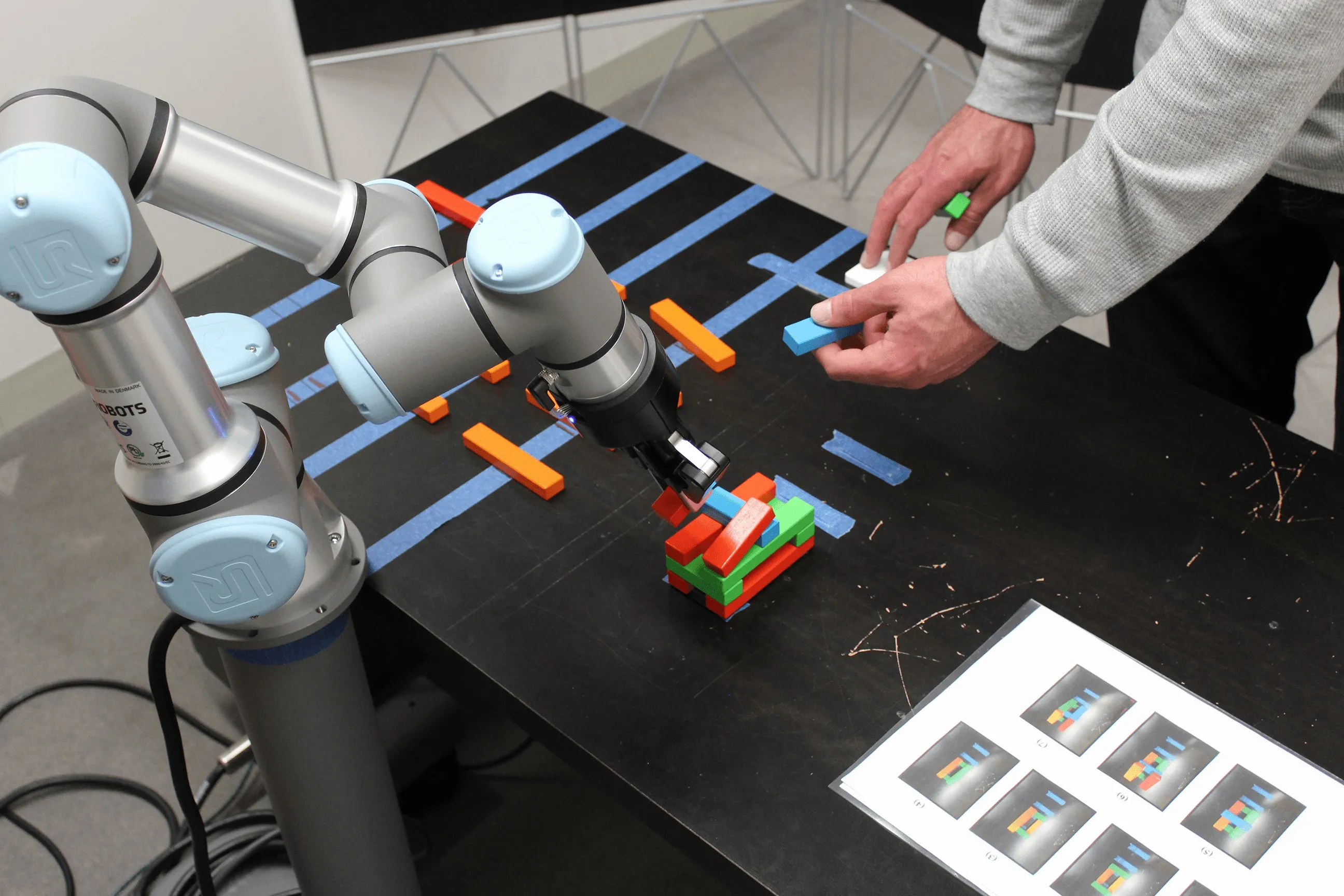 A participant's hands and the UR3e arm building block structures together on a taped-off table, with a sheet of target patterns at the edge.