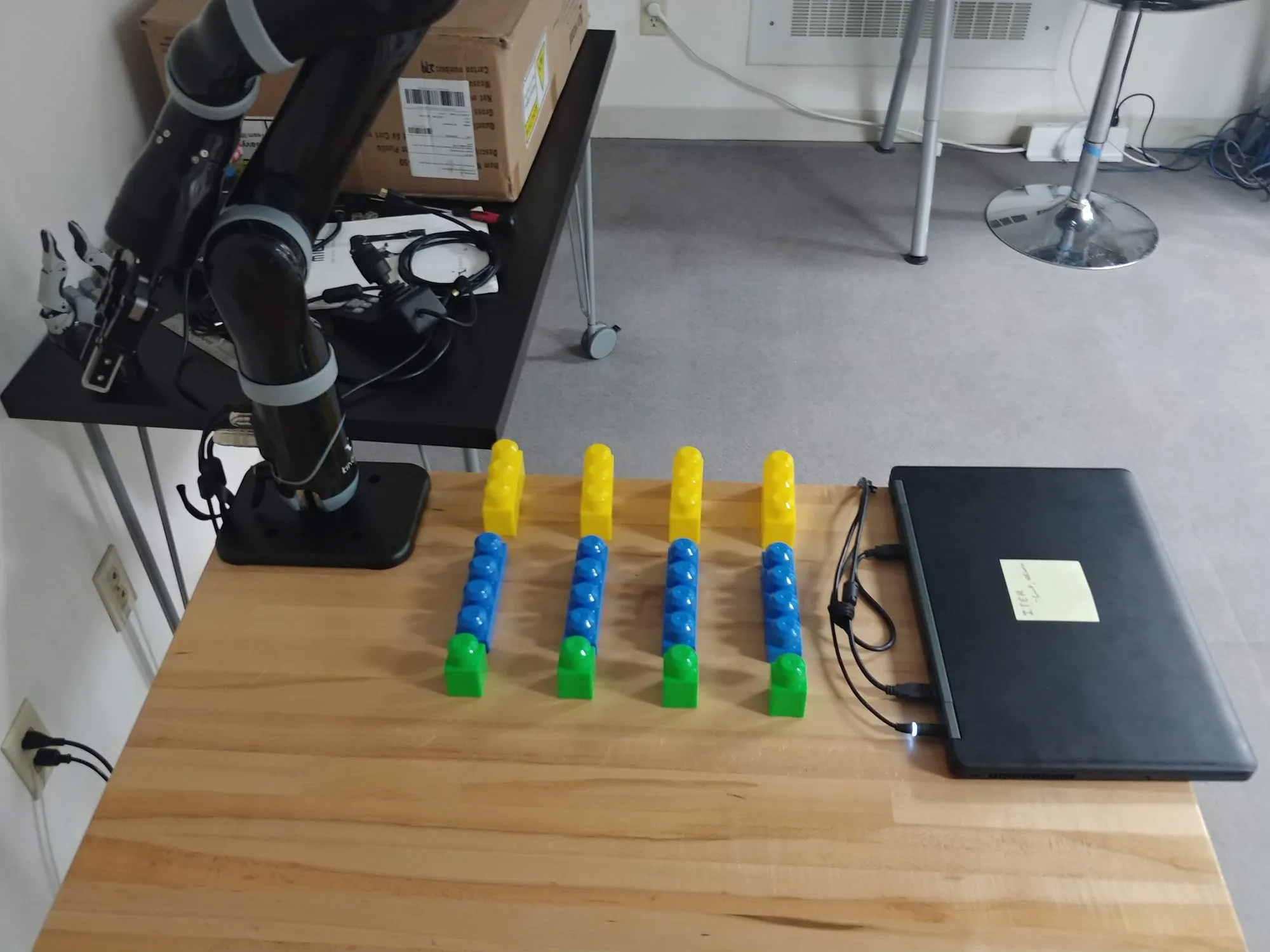 The Kinova at the end of the wooden bench with rows of yellow, blue, and green Mega Bloks laid out in front of it and a laptop to the side, February 2019.