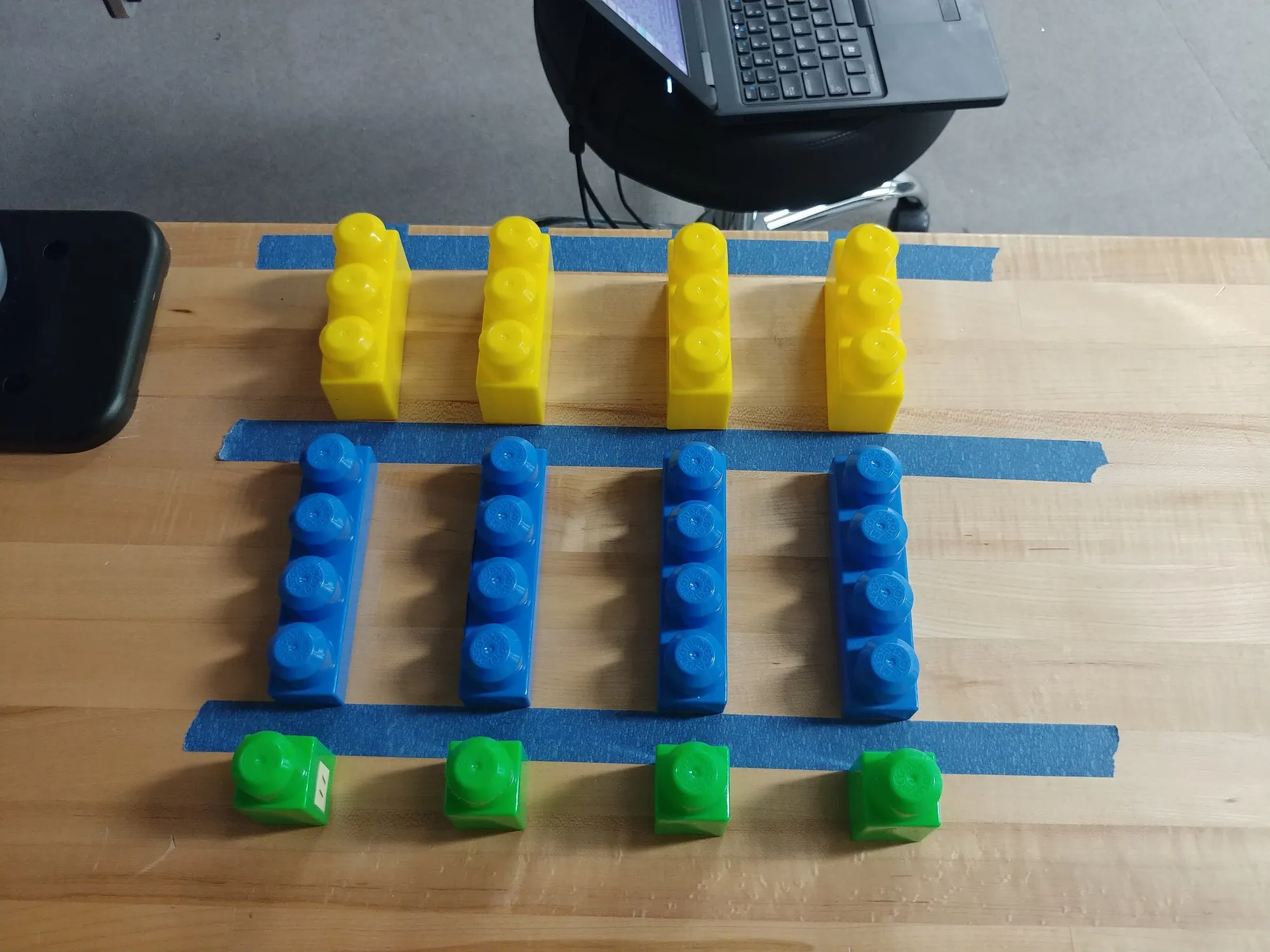The block grid from above: four columns each of yellow, blue, and green blocks on strips of blue tape, a laptop on a cart behind.