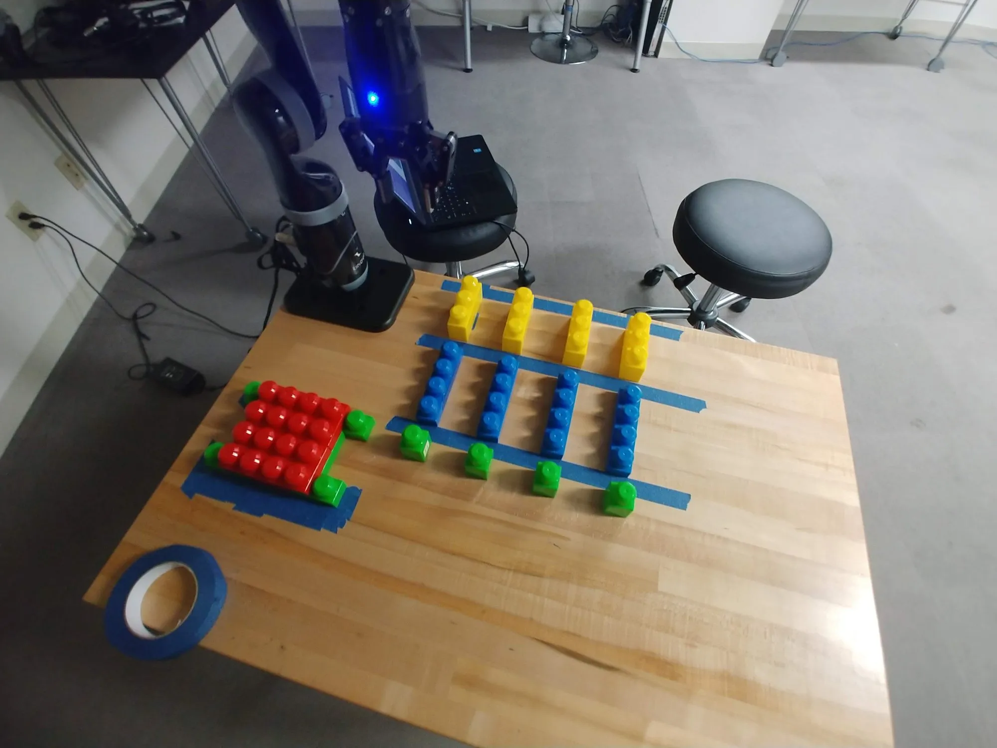The Kinova with its gripper light on, blocks sorted into a red square and color rows on the taped bench, a roll of tape and a stool nearby.