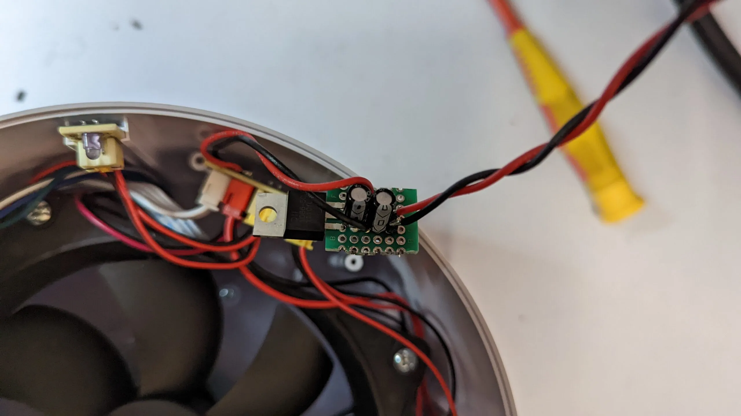 The step-down regulator on a scrap of perfboard, a TO-220 regulator and two capacitors, twisted red and black leads, sitting on the rim next to the fan.