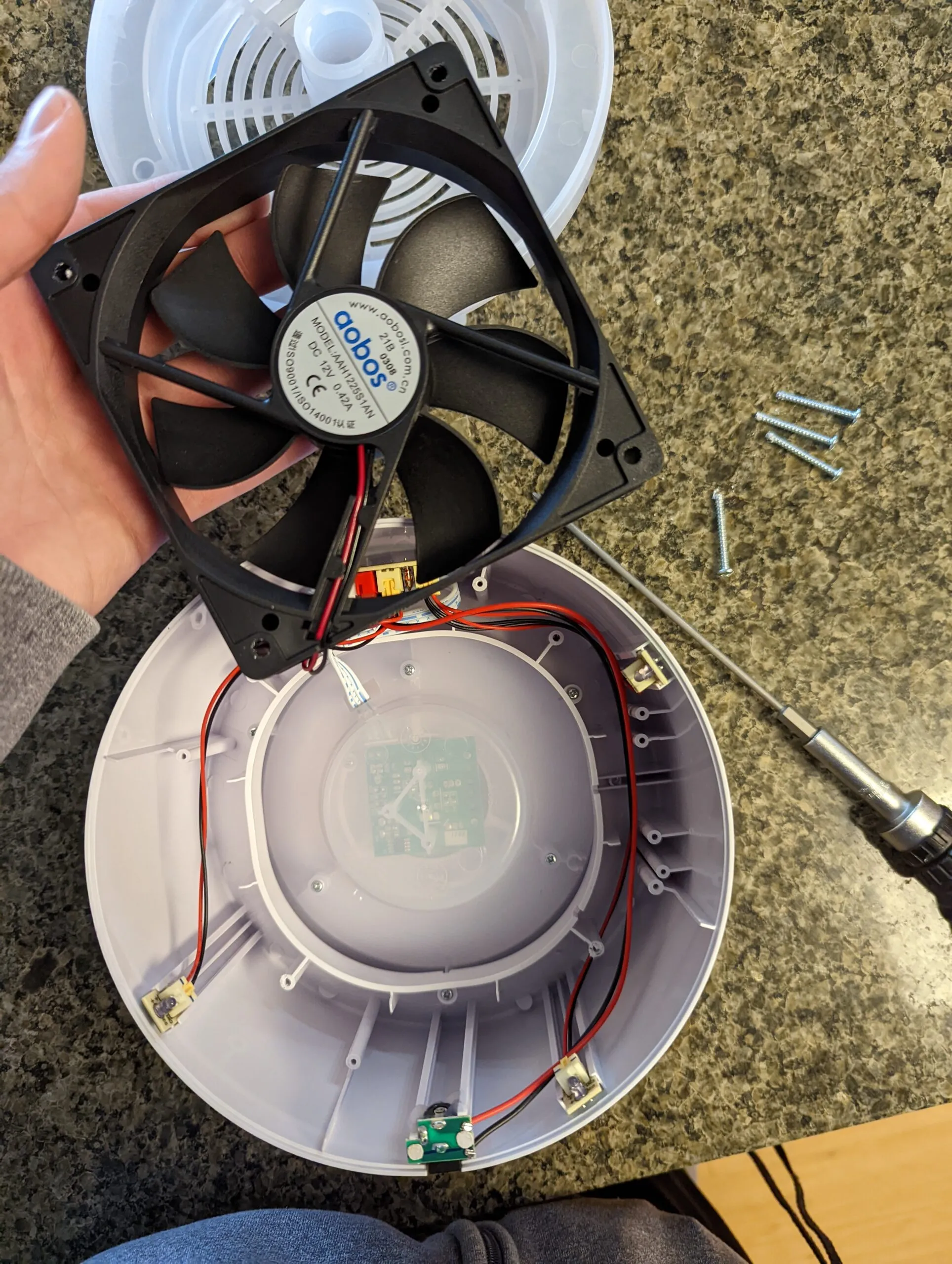 The 12 V Aobos fan freed from the housing, mounting screws scattered on the counter, the control board showing through the translucent center of the shell.
