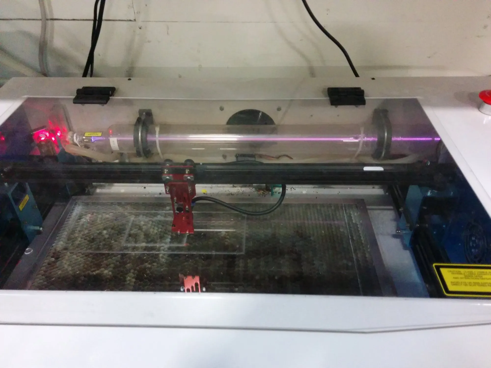 A laser cutter mid-job under its lid, the beam tube along the back and a red-jawed head over the honeycomb bed.