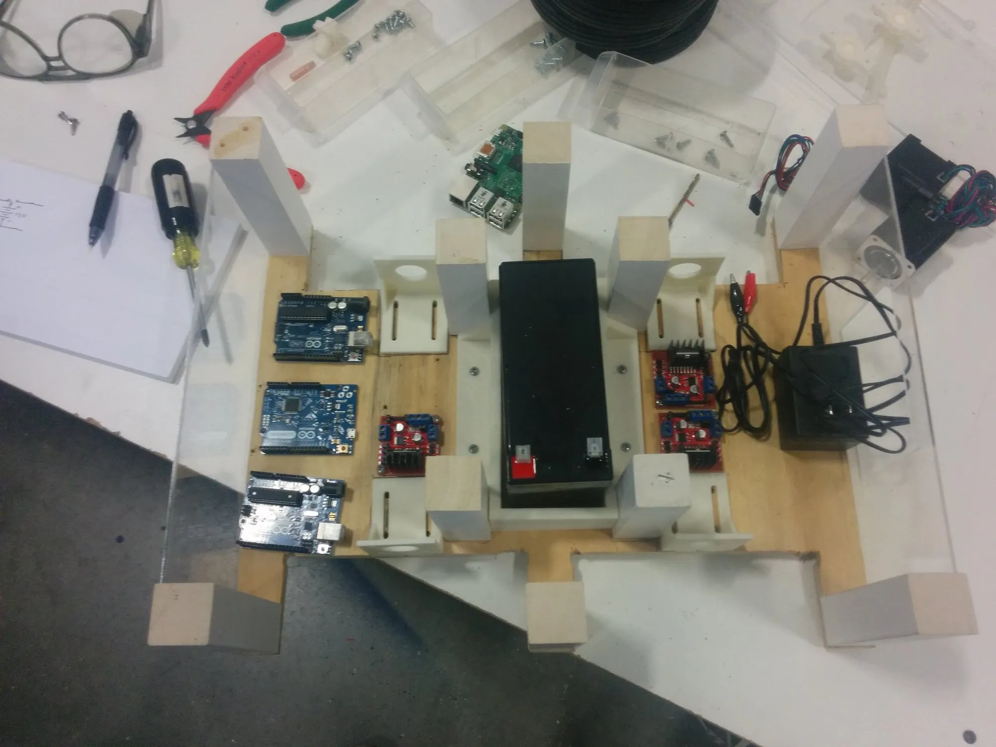 Orion's parts on the bench before assembly: plywood chassis with printed brackets, the SLA battery, a few Arduinos set out for spacing, three of the four motor drivers, the charger, and a Raspberry Pi off to the side.