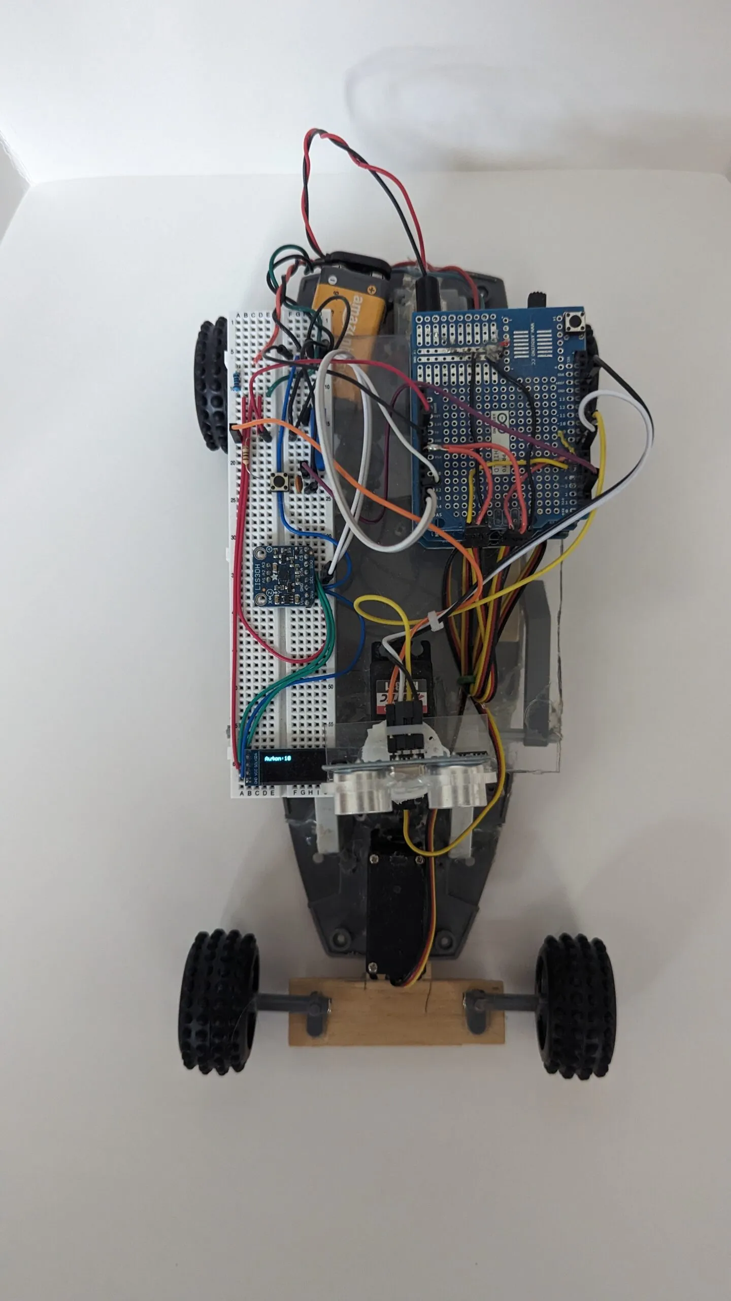 Top-down view of RoverBot: breadboard deck, 9V, and shield stack over the gray chassis, wooden axle at the front.