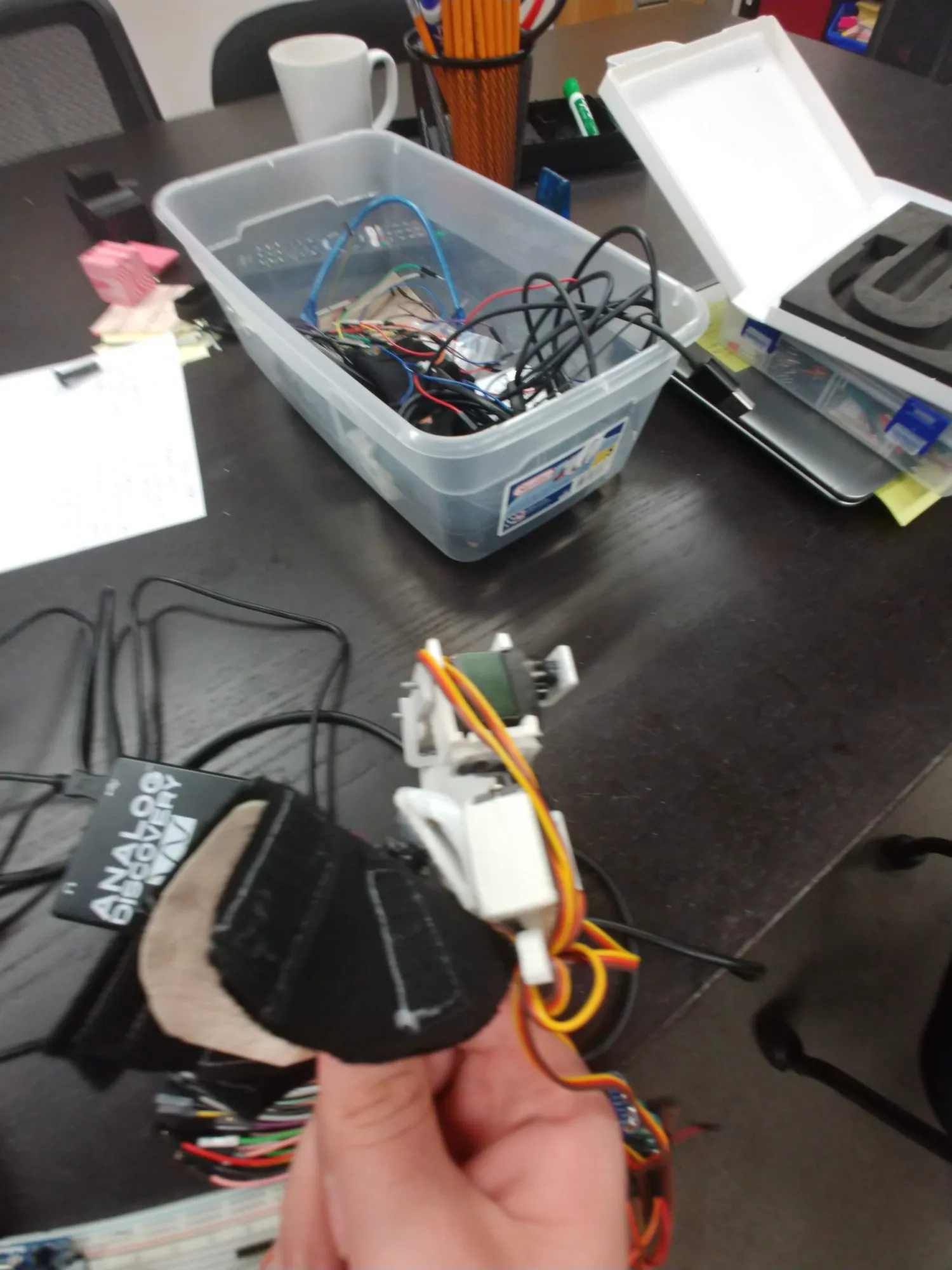 A clear plastic bin of parts and cables on the bench, the finger and its board in the foreground.
