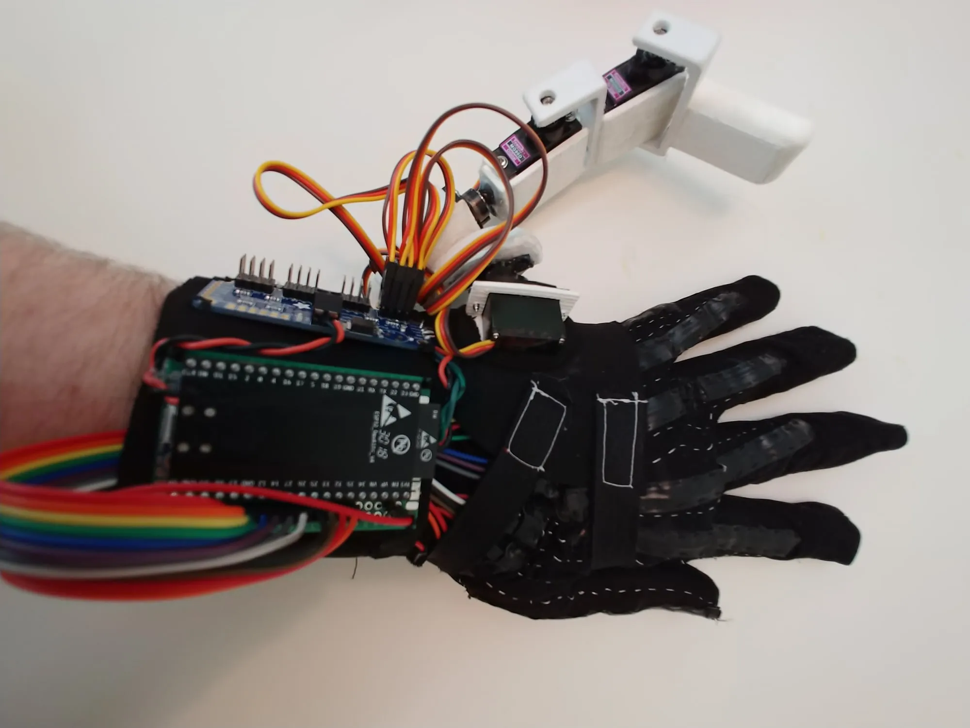 Top-down view of a black glove liner with home-made flex sensors sewn along each finger, an ESP32 dev board and servo driver strapped to the wrist with rainbow ribbon cable, and the 3D printed robotic finger with its servos and orange servo leads mounted beside the little finger.