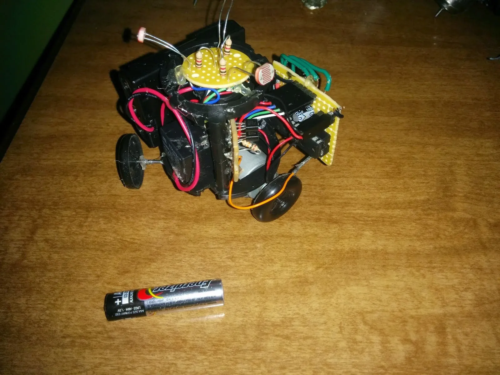 Triclops attempt 1 in 2014: photoresistor sensor disc atop a cylinder body, two of its three wheels visible, AAA battery for scale.