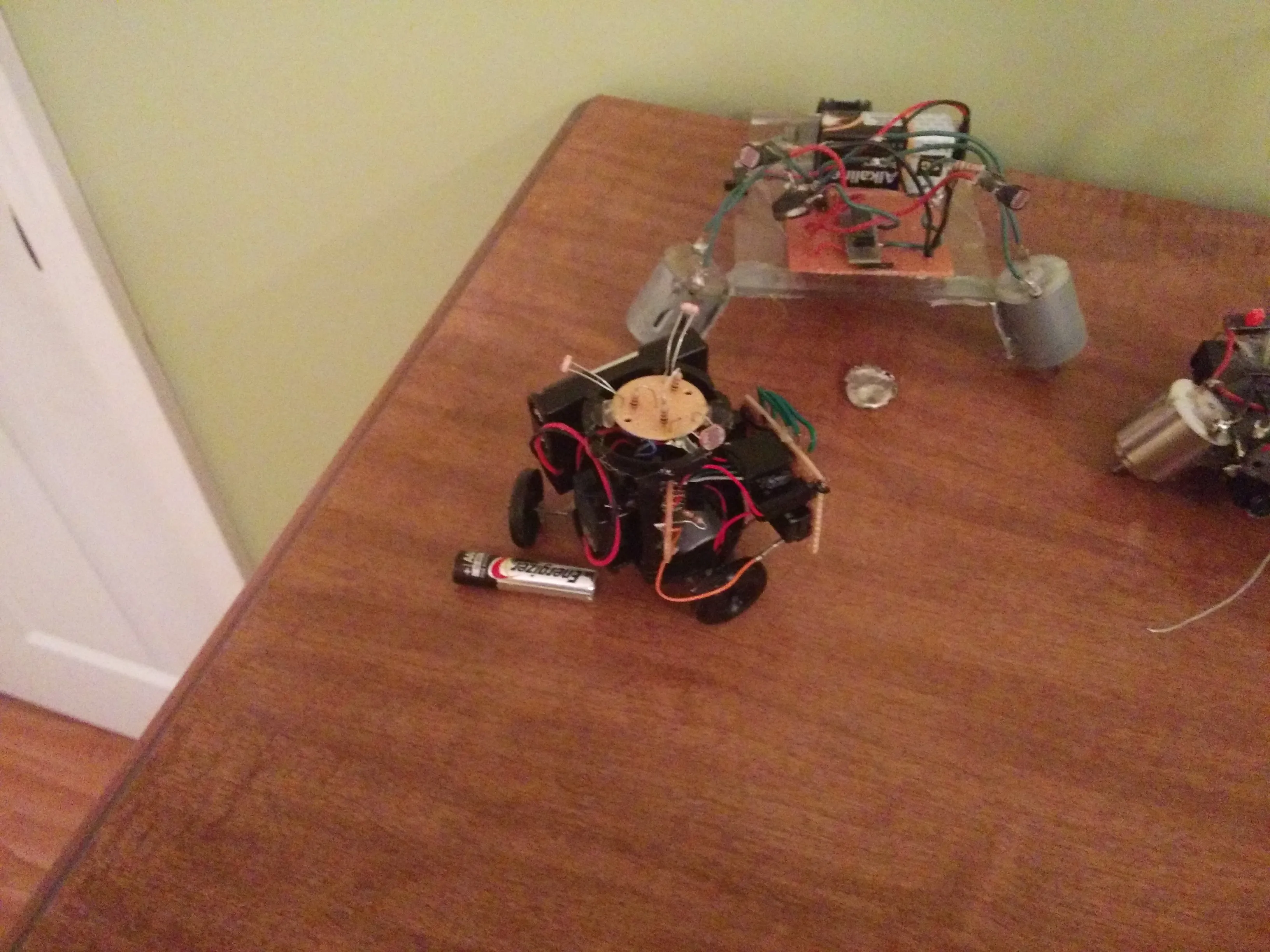 Triclops posed beside an acrylic-framed sibling robot, spare gearmotor at the edge of frame.