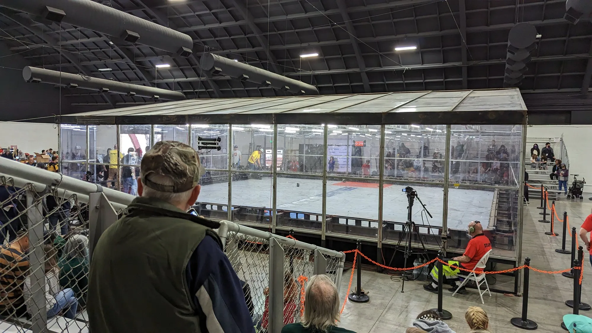 A crowd along a rope barrier watching the polycarbonate heavyweight combat arena in an exhibition hall.