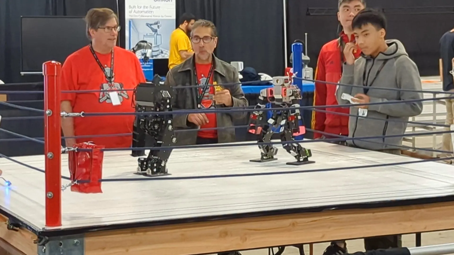 Frame from a video clip: two small humanoid robots facing off in a miniature wrestling ring while their operators stand at the ropes.