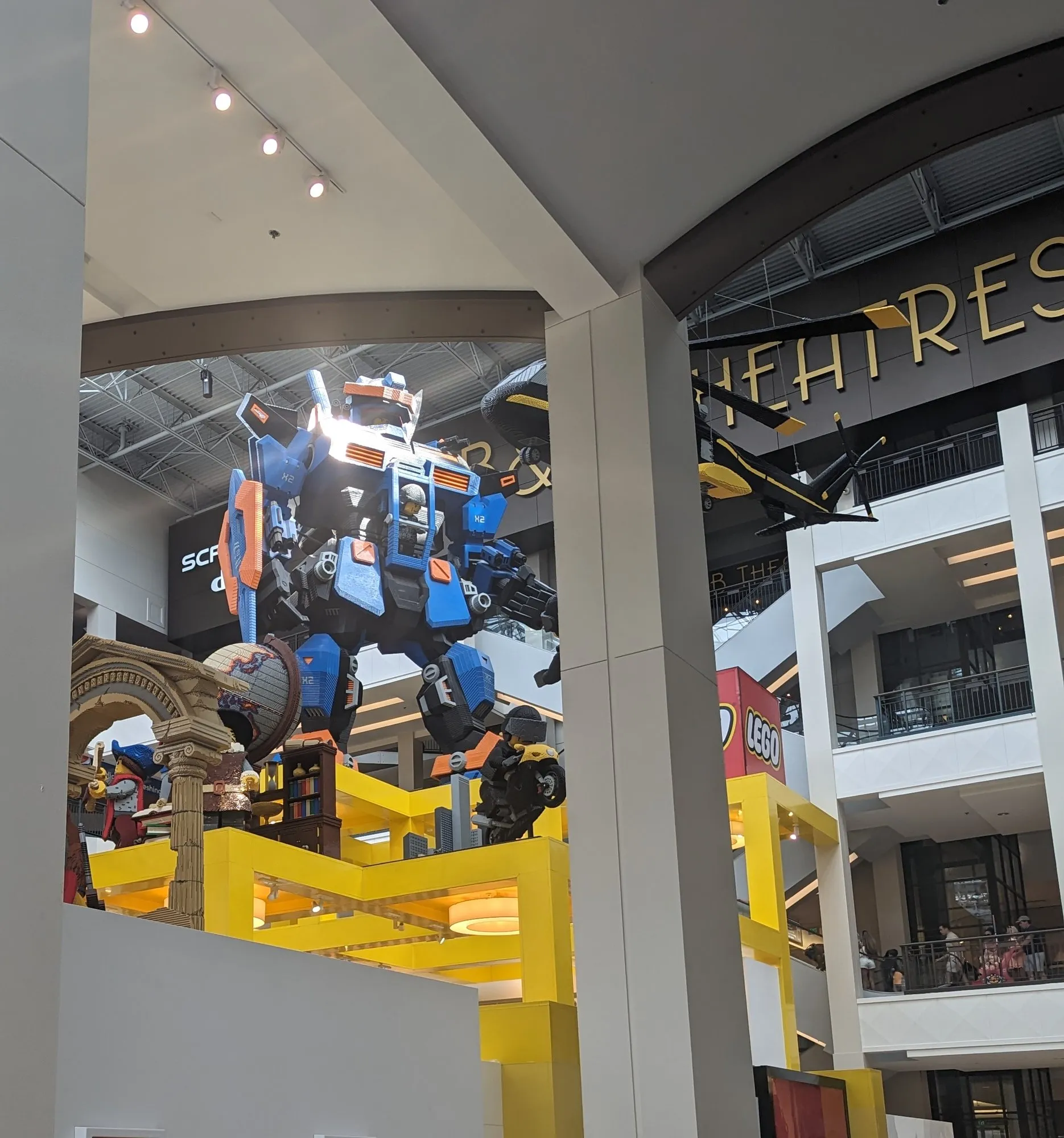 A giant blue and orange LEGO mech standing on top of the LEGO store in a mall atrium, a minifig pilot in its cockpit.