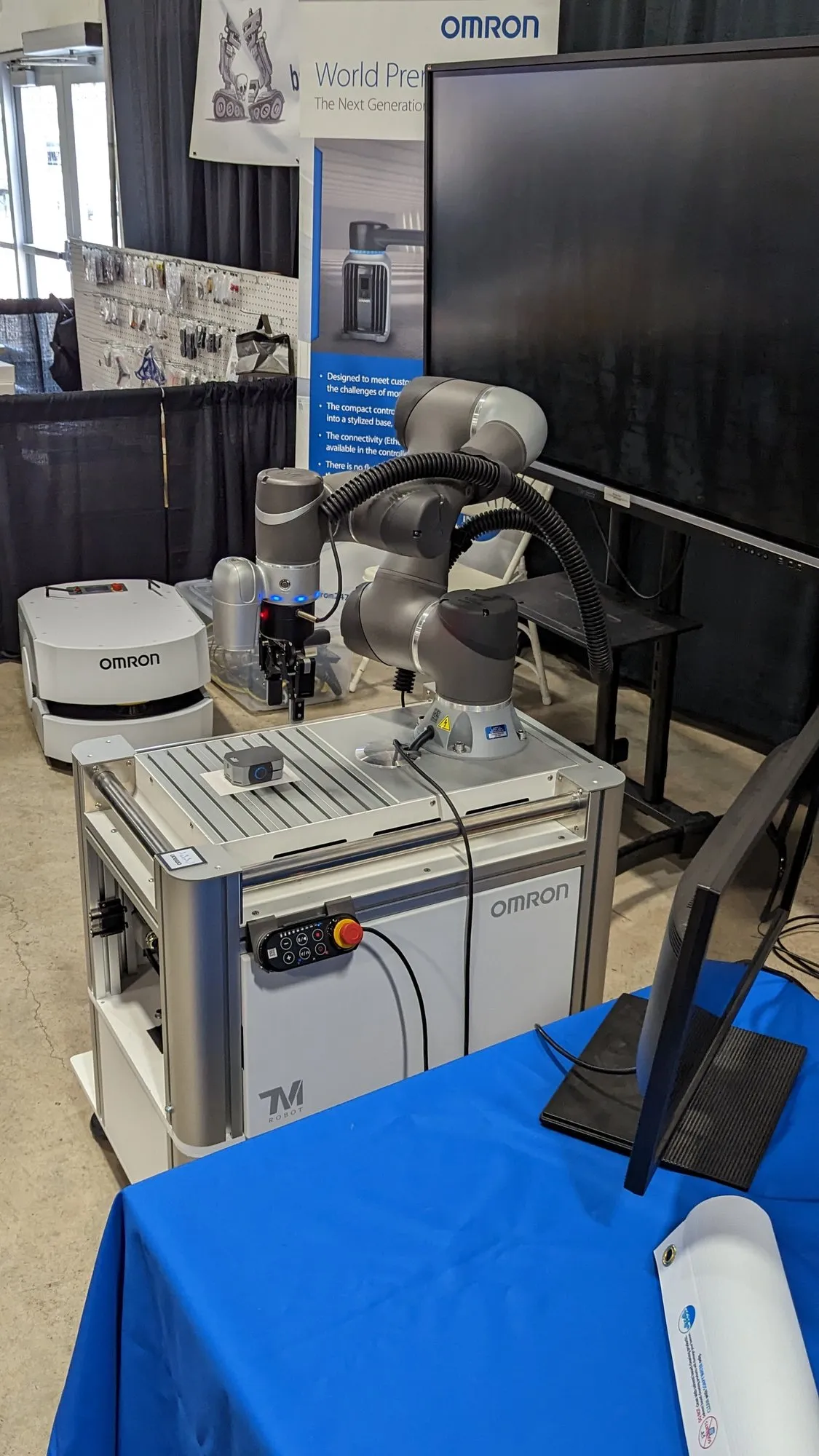 An Omron TM collaborative arm mounted on a mobile cart at a vendor booth, blue tablecloth in front, product banners behind.