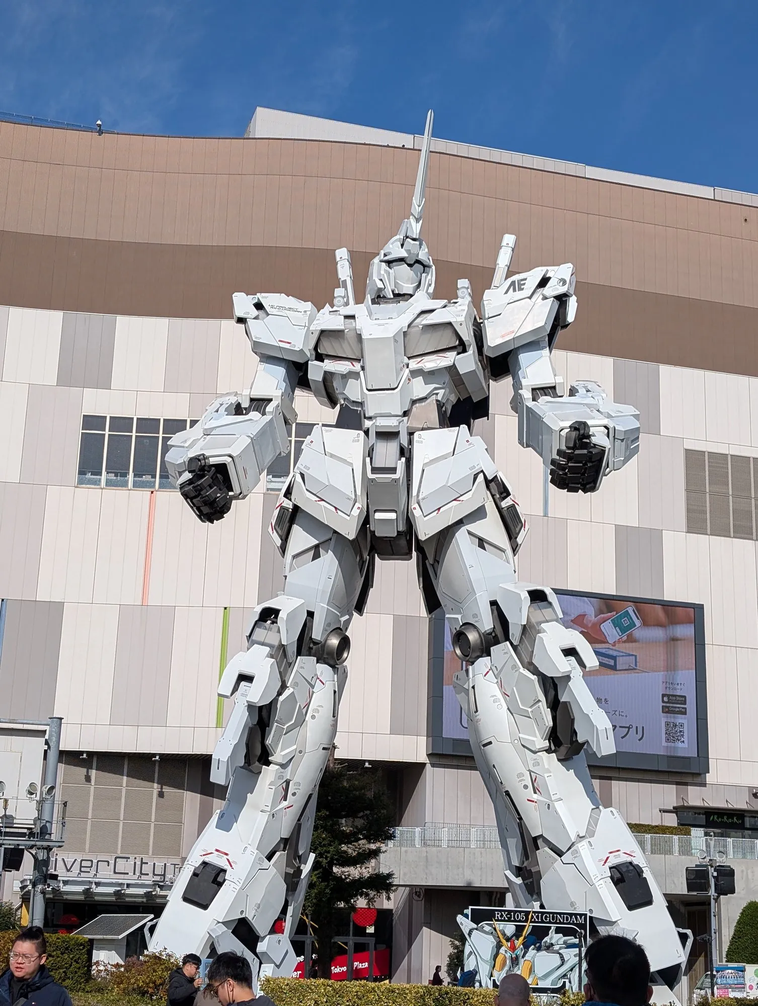 The Unicorn Gundam head-on from the plaza, fists at its sides, DiverCity Tokyo building behind.