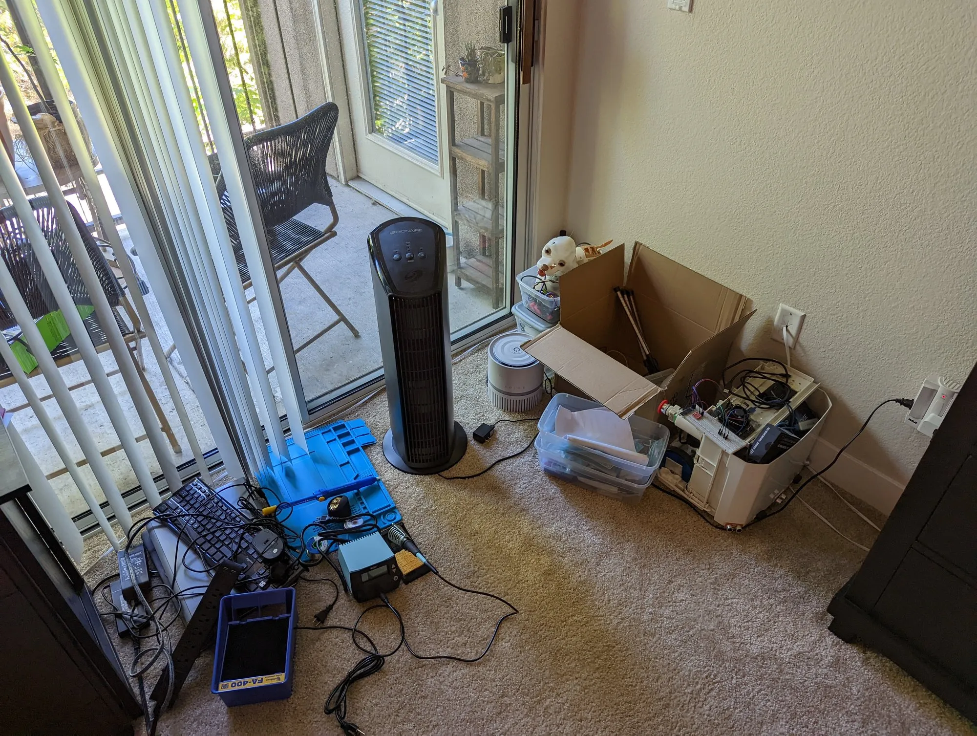 The dehumidifier in the corner of the living room with a breadboard on top, next to boxes and bins of parts; a soldering station, keyboard, and cables on the carpet by the patio door.